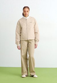 Giacca beige trapuntata con polsini a costine, abbinata a pantaloni beige chiaro. Entrambi i capi hanno una vestibilità comoda. Scarpe semplici a righe completano il look.