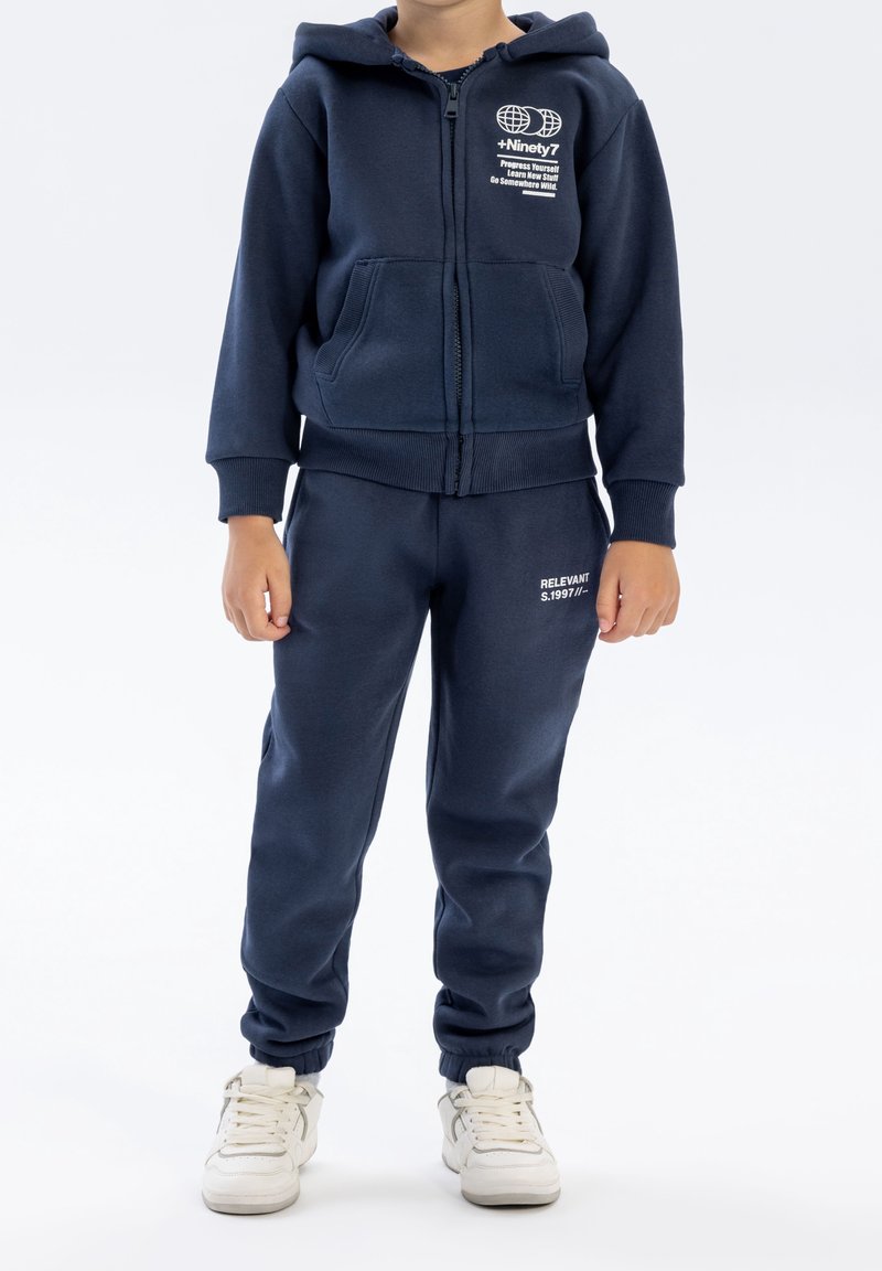 Bambino che indossa una felpa con cappuccio blu navy e pantaloni da jogging abbinati con sneakers bianche, in piedi davanti a uno sfondo bianco uniforme.