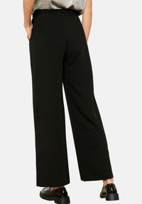 Črne široke hlače z vrvico v pasu. Narejene so iz gladkega materiala z čistimi linijami in diskretnim sijajem, v kombinaciji s črnimi čevlji.