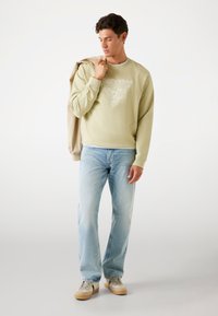Lichtgroene sweatshirt met wit logodesign, gecombineerd met lichtblauwe jeans en beige sneakers. Model houdt een tan jas over één schouder.