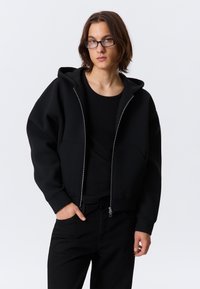 Hanorac negru cu glugă, fermoar integral, două buzunare frontale și manșete striate, asortat cu un top și pantaloni negri mulați. Textură netedă.