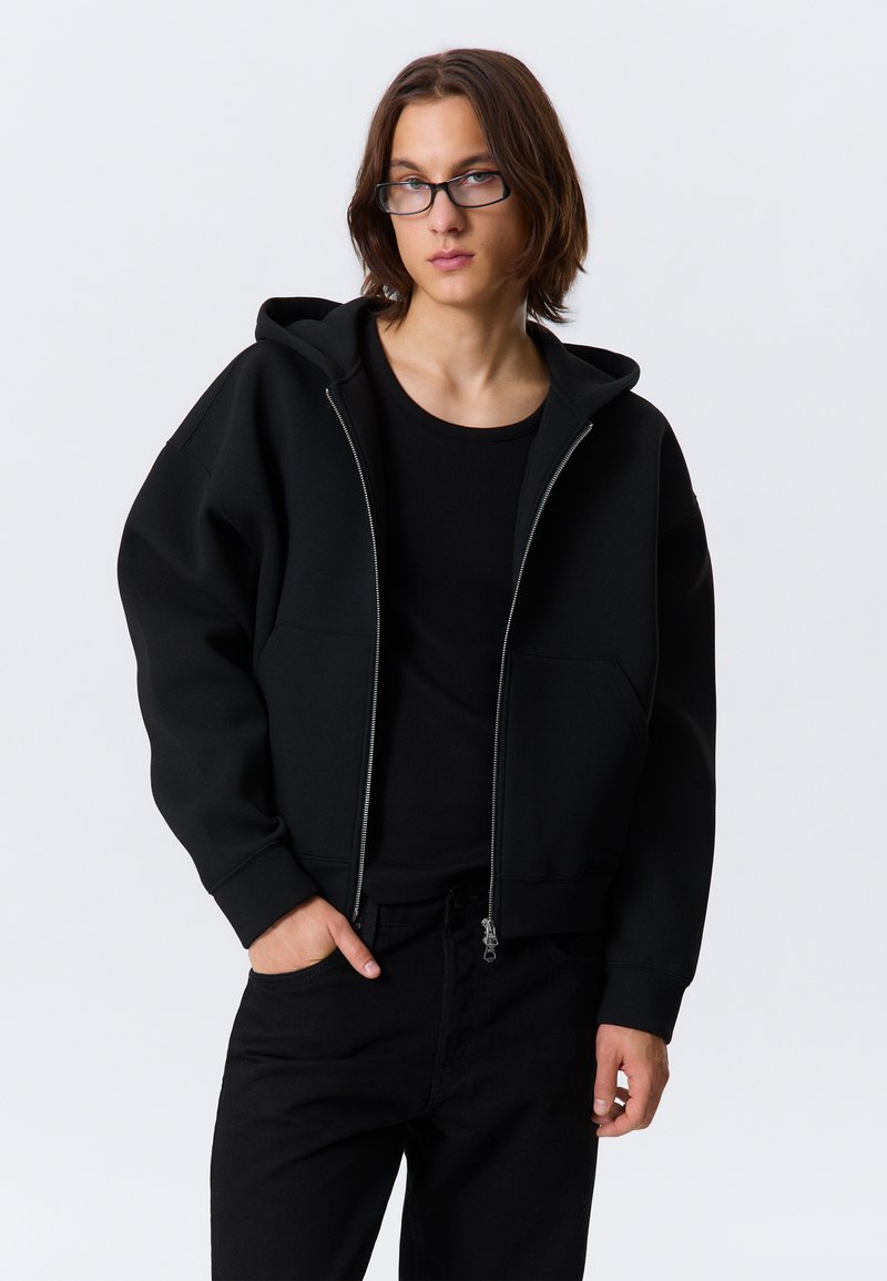 Hanorac negru cu glugă, fermoar integral, două buzunare frontale și manșete striate, asortat cu un top și pantaloni negri mulați. Textură netedă.