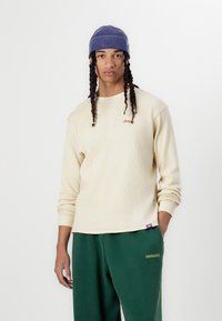 Beige ribbed long-sleeve shirt with a round neckline and embroidered "JACK" detail. Paired with green sweatpants featuring a logo patch.