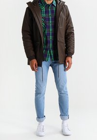 Brązowa kurtka z kapturem nałożona na zieloną i niebieską koszulę w kratę, dopasowana do jasnoniebieskich dżinsów i białych sneakersów. Gładka faktura, luźny fason.