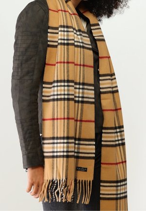 Persona que lleva una bufanda de cuadros beige con rayas negras, blancas y rojas sobre una chaqueta de cuero negra, mostrando los extremos con flecos de la bufanda.