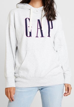 Femme portant un sweat à capuche GAP gris clair avec des lettres bleu marine et un jean bleu clair, debout, les mains relâchées sur les côtés.