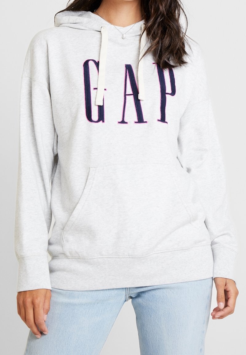 Femme portant un sweat à capuche GAP gris clair avec des lettres bleu marine et un jean bleu clair, debout, les mains relâchées sur les côtés.
