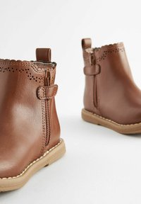 Bottines en cuir marron avec un haut à bords festonnés, des zips latéraux et des semelles en caoutchouc beige. Perforations décoratives près du haut.