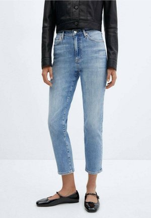 Kvinde iført lyseblå korte jeans, sort cropped læderjakke og sorte Mary Jane flade sko står foran en ensfarvet baggrund.