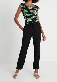 Zwarte elastische broek met een fitted top met een bloemenpatroon in levendige kleuren en doorschijnende mouwen. Sandalen maken de look compleet.