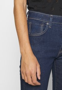 Mörkblå denimjeans med en slät textur, kontrasterande gyllene sömmar och en detalj på framfickan, visade med en hand som vilar på midjebandet.