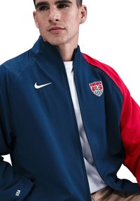 Blaue und rote Nike-Jacke mit einem Reißverschluss, elastischen Bündchen und USA-Emblem auf der linken Brust. Aus glattem Material gefertigt, lockere Passform.