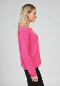 Pull en tricot rose à coupe décontractée, texture côtelée, encolure ronde et épaules tombantes. Porté avec un pantalon noir. Accessoire : bague au doigt.