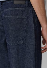 Ciemnoniebieskie jeansy denimowe o prostym kroju, z widoczną tylną kieszenią i skórzaną naszywką z logo na pasku. Gładka faktura.