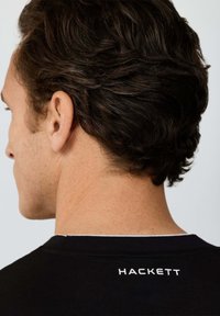 Mężczyzna o krótkich kręconych ciemnych włosach, widziany od tyłu, ubrany w czarną koszulę z nadrukiem "HACKETT" na tylnej części kołnierza.