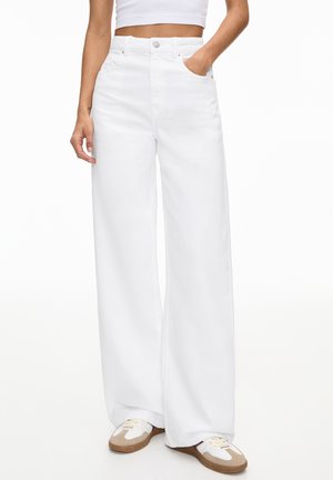 Kvinde iført hvide højttaljede vide jeans, kort hvid top og beige-hvide sneakers med den ene hånd i lommen.