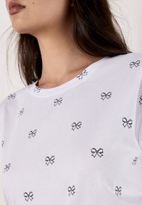 Un primer plano de la parte superior del cuerpo de una mujer que lleva una camiseta blanca decorada con pequeños motivos de lazo negro, mostrando un aspecto casual y elegante.