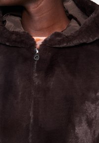 Dunkelbraune Kunstpelz-Zip-Jacke mit weicher Textur, ausgestattet mit einer Kapuze und einem Reißverschluss. Gefüttert mit einem kontrastierenden glatten Stoff.