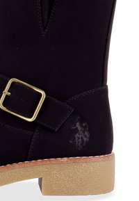 Partie de botte noire avec sangle à boucle dorée, semelle beige, et logo Polo embossé représentant deux joueurs de polo à cheval.