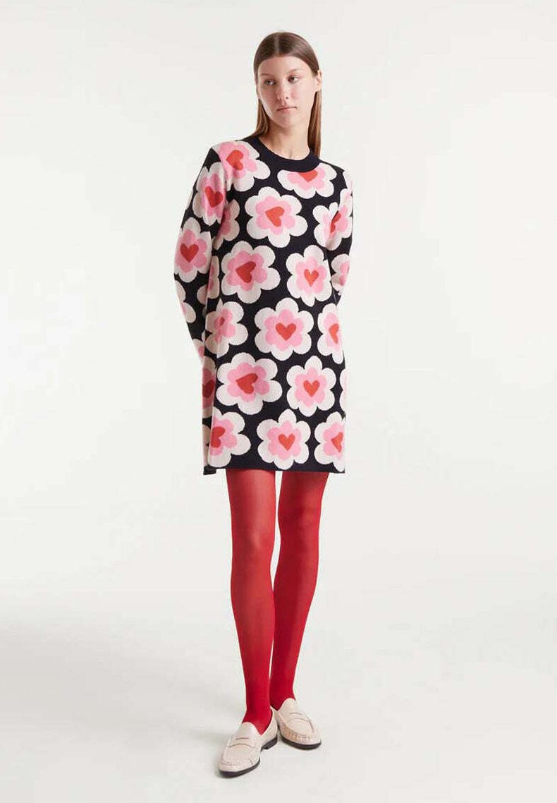 Mujer vestida con un vestido negro con grandes flores rosas y rojas, medias rojas y mocasines beige, de pie con las manos detrás de la espalda sobre un fondo blanco.