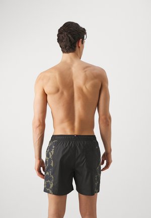 Bañador negro con acentos de camuflaje verde en los laterales, cintura elástica y bolsillo trasero. Modelo mostrado de espaldas.