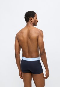 Boxer briefs bleu marine avec une ceinture élastique bleu clair portant le logo Lacoste, texture côtelée, et coupe ajustée sur un mannequin masculin.