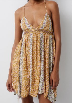 Frau trägt ein ärmelloses Sundress mit V-Ausschnitt und dünnen Trägern, das ein Blumenmuster in Senfgelb, Weiß und Rosatönen aufweist.