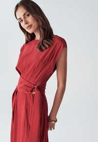 Červené šaty po kolena s hladkým povrchem, s přitaženým pasem a zlatým prstencem jako akcentem. Krátké, široké rukávy.