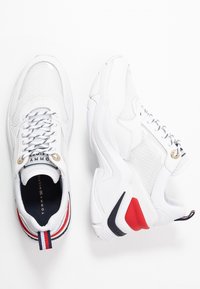 Baskets de sport blanches avec une tige en mesh, œillets dorés et un accent rouge et noir sur la semelle. Les lacets sont noirs avec un motif blanc.