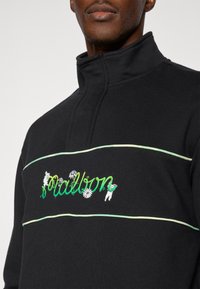 Μαύρο φούτερ με ψηλό γιακά, πράσινη και λευκή κεντητική με λουλουδένιες λεπτομέρειες, και αντίθετο οριζόντιο μοτίβο γραμμών.