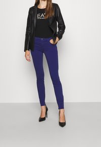 Blazer en cuir noir sur une chemise noire avec texte ; jeans violet ajustés ; talons hauts noirs ; textures lisses, design moderne, coupe sur mesure.