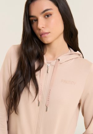 Jeune femme aux longs cheveux foncés portant un sweat à capuche zippé beige clair avec une capuche et le logo "FREDDY" sur la poitrine.