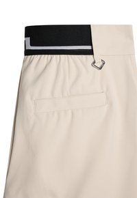 Beige housut, joissa on musta joustava vyötärö, jossa on hopeinen D-ring, ja takatasku, jossa on ommeltu sulku. Sileä kankaan rakenne.