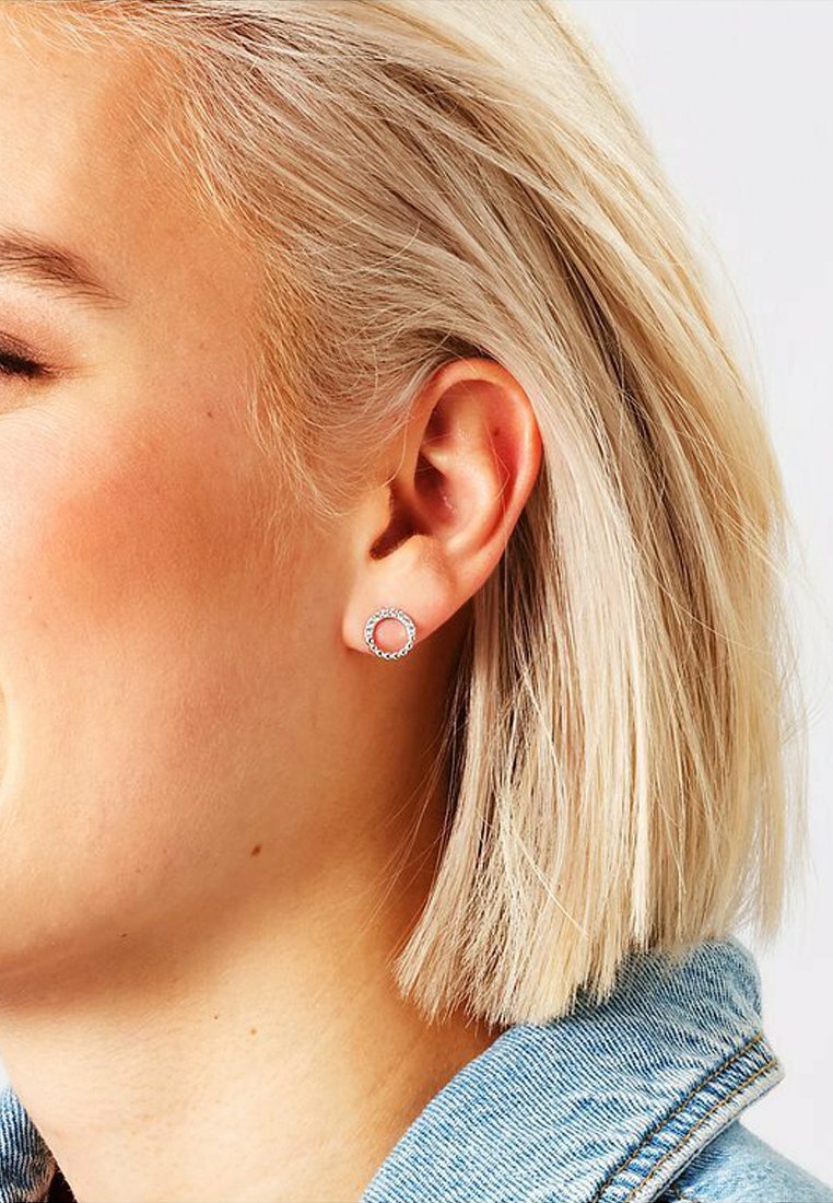 Silverfärgad cirkulär stikörhänge med små klara ädelstenar, med en texturerad yta och en polerad finish, båd på en kort blond frisyr.