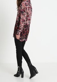 Durchsichtige Bluse mit langen Ärmeln und floralem Muster in Rosa- und dunklen Tönen, getragen über schwarzen figurbetonten Hosen und schwarzen Absatzstiefeletten.