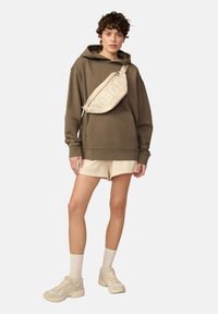 Oliwkowy hoodie z kieszenią na przodzie, w połączeniu z beżowymi szortami i jasnobeżową torbą na ramię z wyrazistym napisem. Jasne sneakersy i skarpetki.