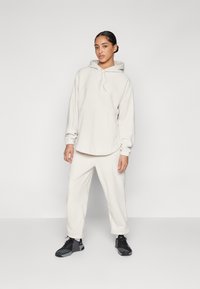 Felpa beige chiaro con cappuccio e lacci, vestibilità oversize, polsini a coste, abbinata a pantaloni della tuta a sigaretta coordinati. Scarpe da ginnastica nere completano l'outfit.