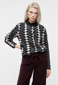 Ilgaplaukė moteris su ilgaa plaukų sukasi, vilkinti juodai ir smėlio spalvos rombų megztinį, virš juodo marškinėlio su apykakle ir bordo spalvos aksominius kelnes.