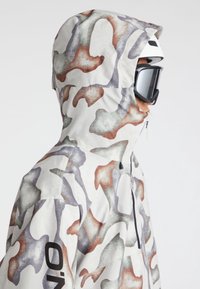 Skijacke in Creme mit einem mehrfarbigen, abstrakten Camouflage-Muster in Grautönen, Braun und Weiß; mit Reißverschluss und Kapuze.