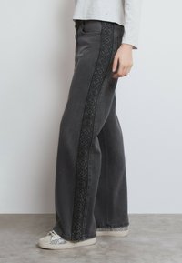 Persona con pantalones vaqueros negros de pierna ancha con paneles laterales bordados y zapatillas blancas con detalles de purpurina plateada, de pie sobre un suelo gris.