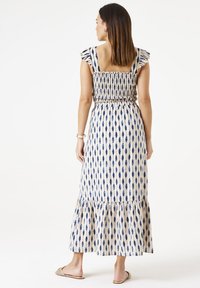 Maxikleid in Creme mit blauem Tropfenmuster, gerafftem Oberteil, RüSCHENTRÄGERN und geschichteten Saum. Trägt flache Sandalen.