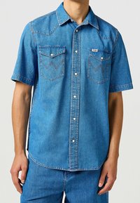 Kortärmad denimskjorta i mediumblått, med två bröstfickor, spetsig yoke, tryckknappar och kontrasterande sömdetaljer.