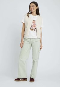 Gilet en polaire beige avec deux poches avant, porté sur un t-shirt graphique blanc avec un motif floral coloré, associé à un pantalon vert clair et des chaussures bordeaux.