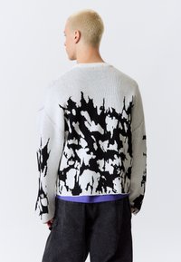 Pull en tricot avec une base grise et un motif abstrait noir contrastant, col rond et coupe décontractée, associé à une chemise superposée.