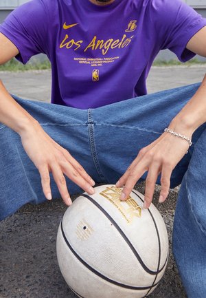 Person, der bærer en lilla Los Angeles Lakers-trøje og blå jeans, holder en hvid basketball, mens de sidder på asfalt.
