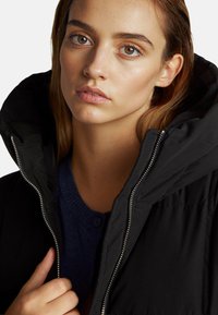 Czarna kurtka puchowa z wysokim kołnierzem i srebrnym zamkiem błyskawicznym. Noszona na granatowym swetrze, podkreślająca miękkie tekstury i warstwowość.