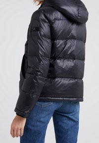 Person iført en sort quiltet dunjakke med hætte og blå jeans, vist bagfra mod en ensartet lys baggrund.