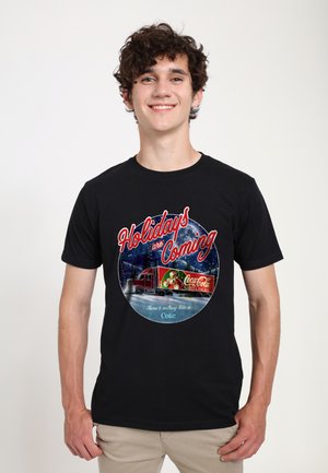 Black t-shirt featuring a round graphic with a red Coca-Cola truck, Christmas theme, snowy background, and white text reading “Holidays are Coming.”