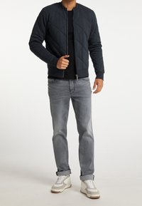 Vadderad marinblå bomberjacka med rund halsringning, tillsammans med en svart skjorta, ljusgrå jeans och vita sneakers.