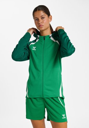 Vrouw die de capuchon van een groene Hummel sportjas rechtzet, met bijpassende groene korte broek met witte accenten, staand tegen een witte achtergrond.